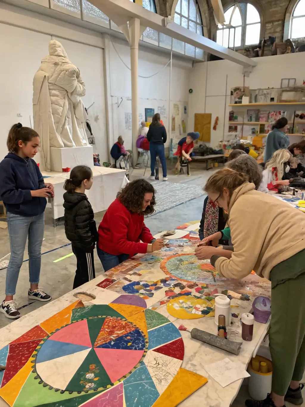 A group of children and adults engaged in a hands-on art workshop, painting and sculpting together in a bright community hall, with completed artworks displayed on the walls.